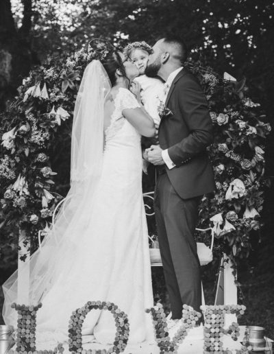 Tendre étreinte : les mariés embrassent leur fille sur la joue lors de la cérémonie laïque sous une arche de fleurs, à leurs pieds le mot 'love'.
