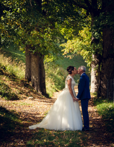 Étreinte automnale : Les mariés s'embrassent tendrement, main dans la main, sous l'ombrage d'une allée d'arbres aux couleurs automnales.