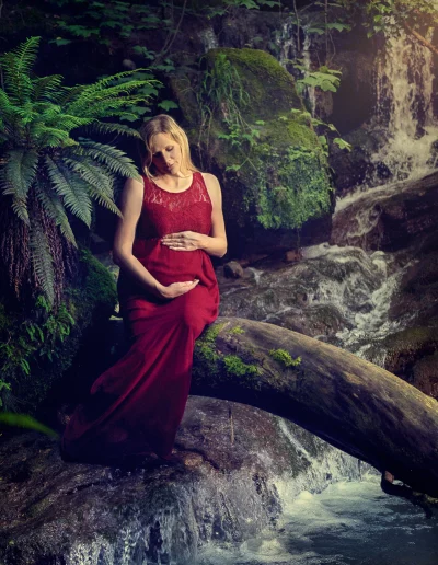Femme enceinte épanouie, assise sur un tronc au-dessus d'une cascade lors d'une séance photo grossesse en pleine nature