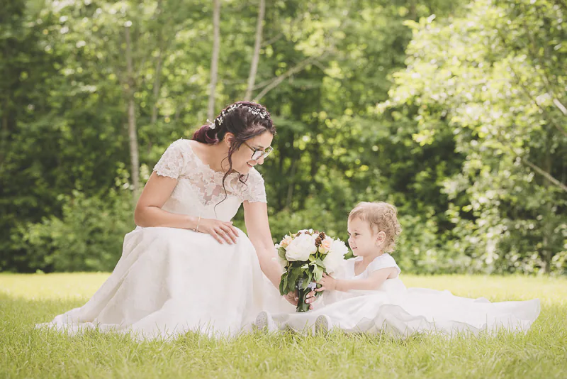 Une mariée élégante tient son bouquet de fleurs blanches tandis que sa fille sourit à ses côtés, toutes deux sur une pelouse verte