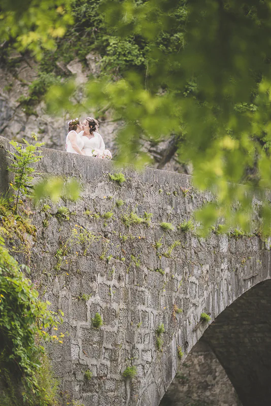 Baiser passionné entre deux femmes sur un pont en pierre, Cluses, Haute-Savoie. Capturé à travers les feuillages d'un arbre.