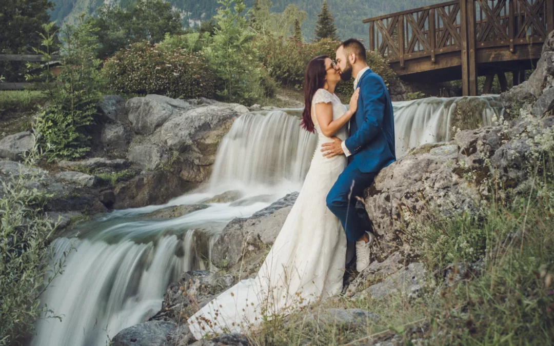 Mariés s'embrassant tendrement au bord du lac à Samoëns, avec une cascade en arrière-plan et une montagne majestueuse au loin.