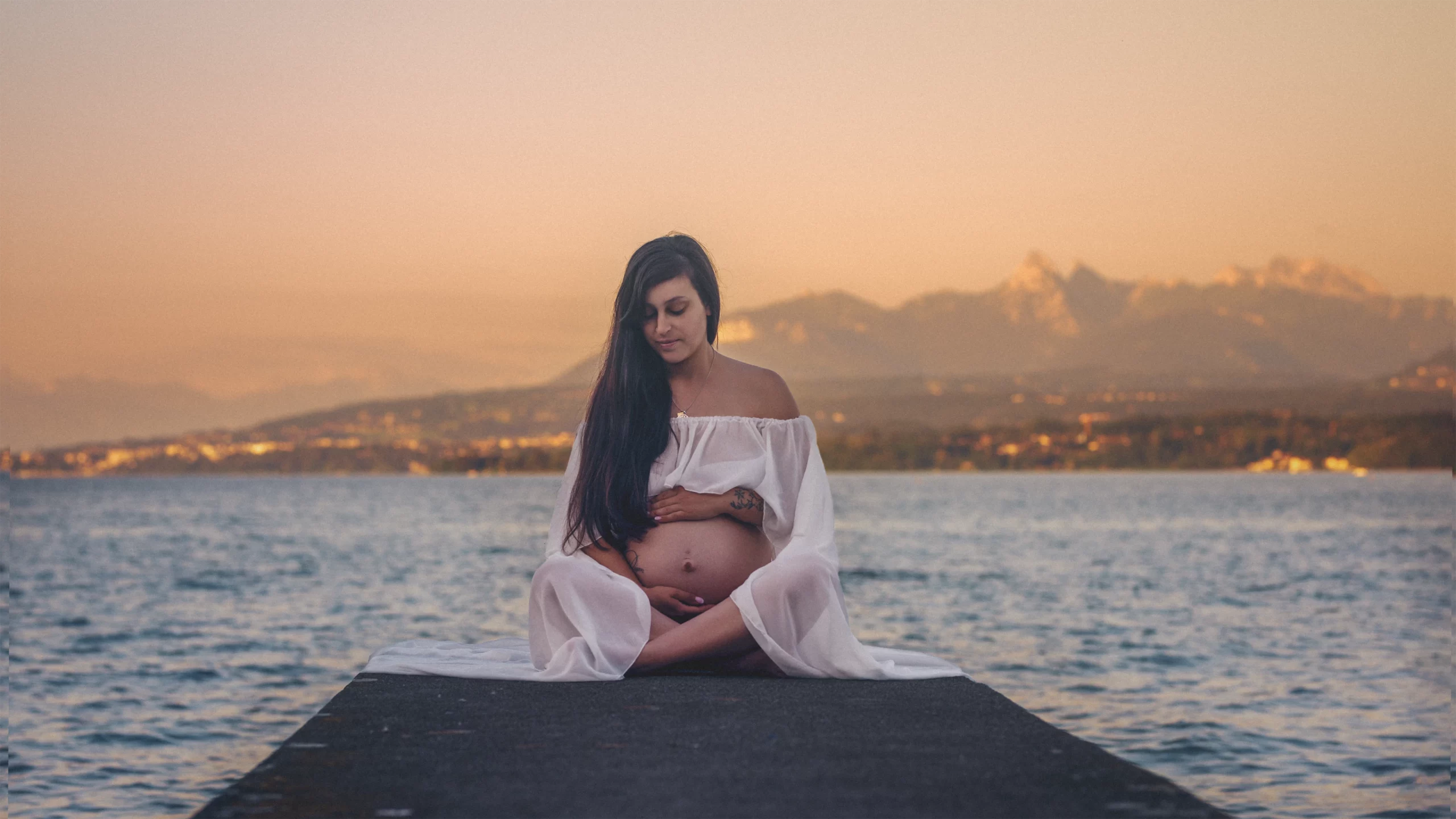 Grossesse au bord du Léman : Un moment unique à Thonon Séance photo de maternité à Thonon : gros plan sur une future maman assise sur un ponton, le lac et les montagnes visibles à l'arrière.