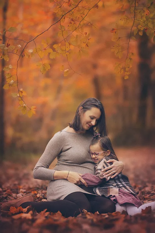 Séance photo de famille en Haute-Savoie en automne : Câlin entre une maman et sa fille Une maman et sa petite fille assises sur un tas de feuilles mortes en automne, la petite fille blottie contre sa maman pour un câlin, lors d'une séance photo de famille en Haute-Savoie.